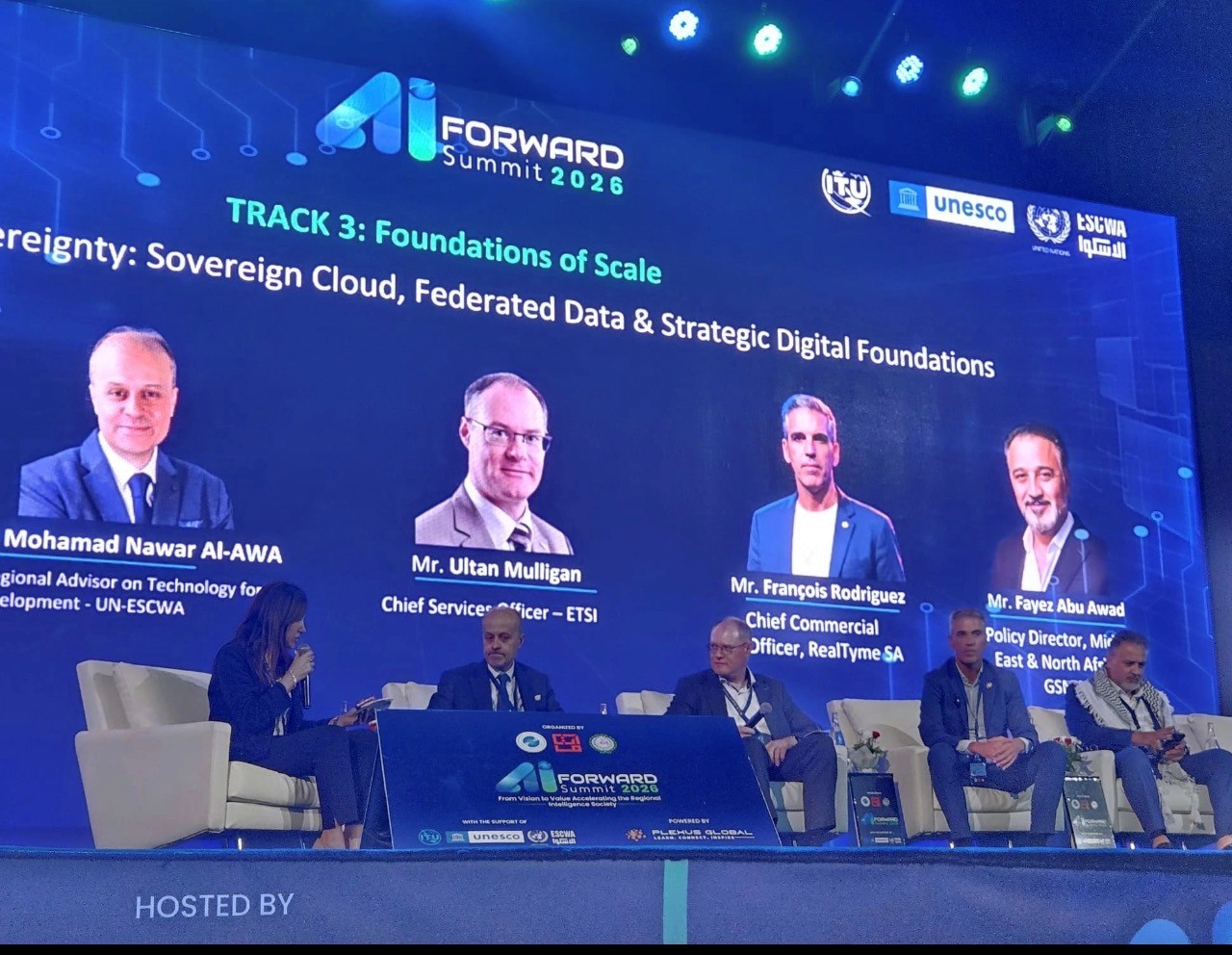 Panel of speakers, seated, with screen showing behind them