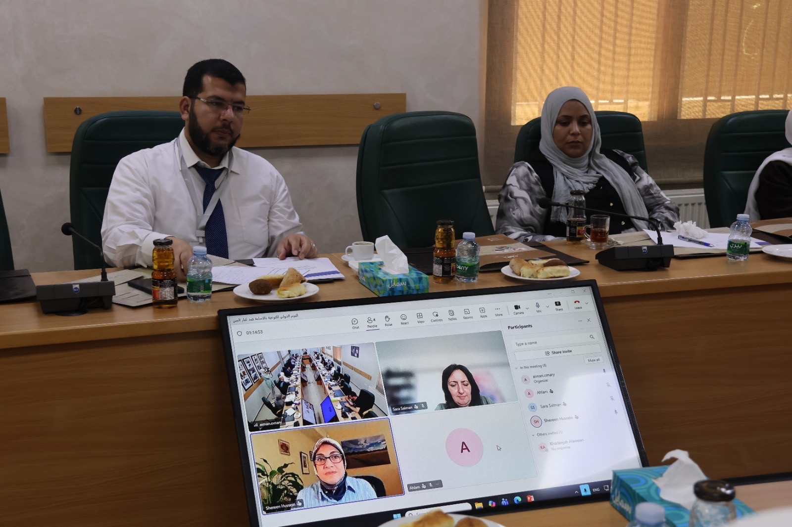 Two participants behind the table, with a screen showing online participants